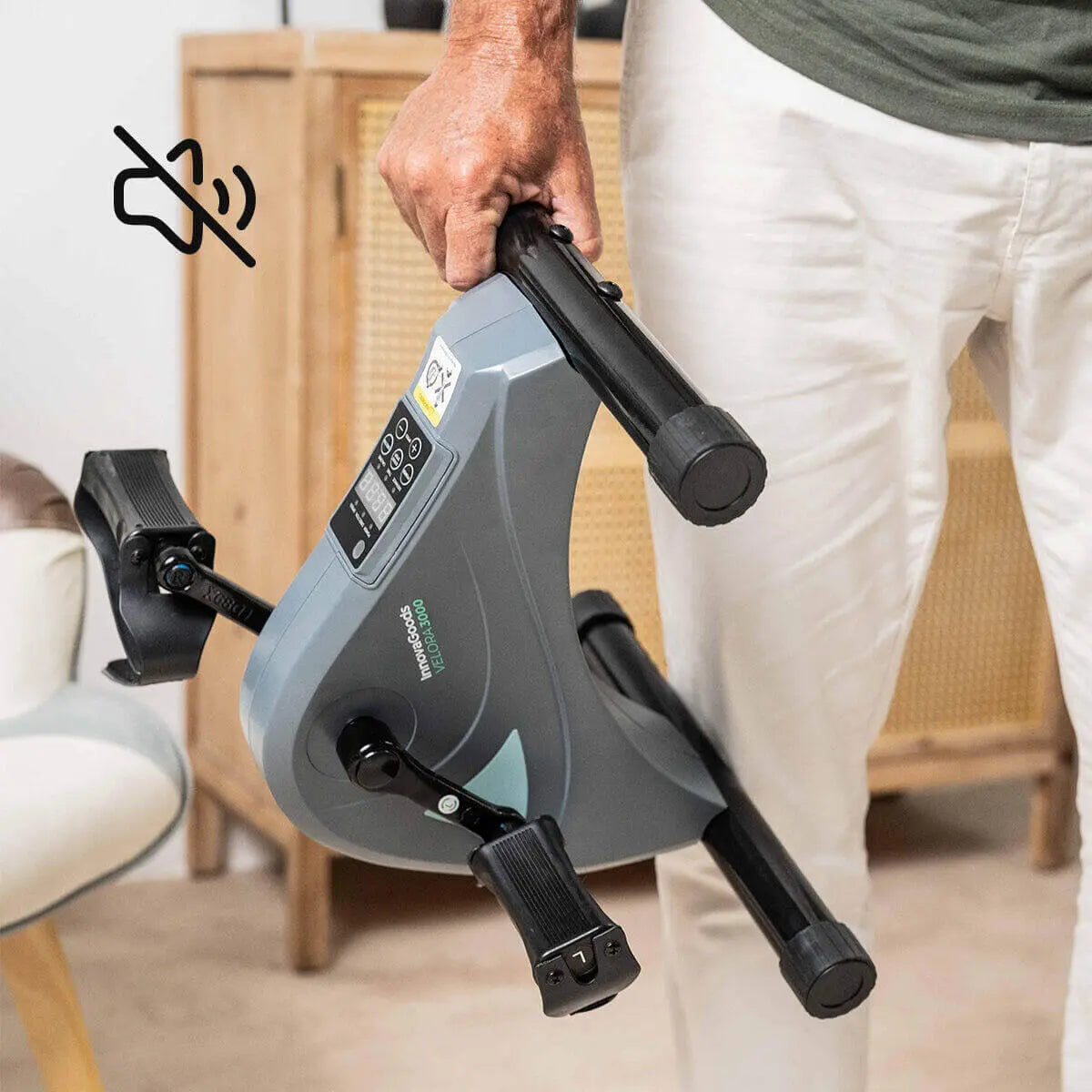 Persona sosteniendo un pedaleador eléctrico gris compacto para brazos y piernas con pantalla y pedales negros.