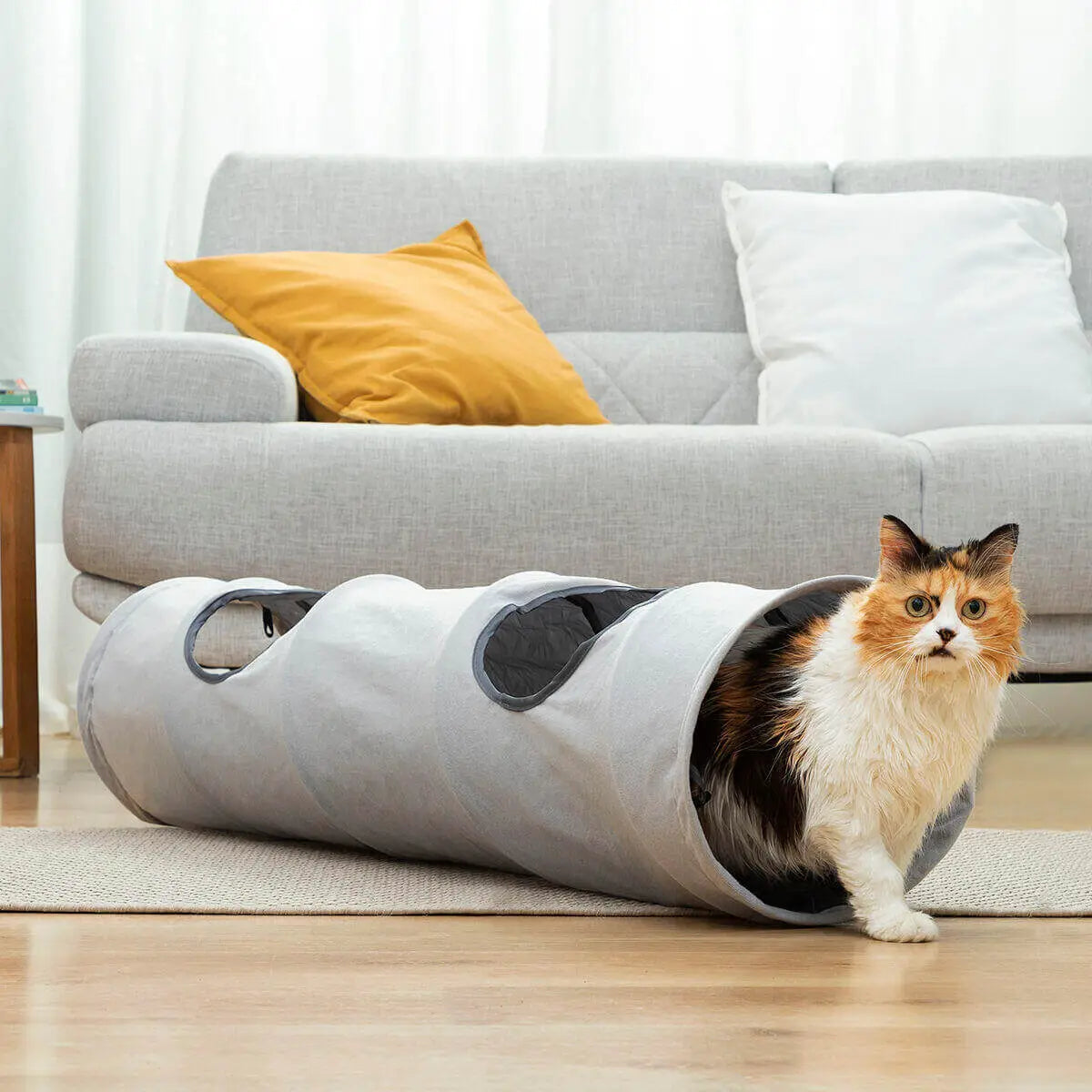 Gato tricolor saliendo de un túnel de juego gris plegable con aberturas laterales, ubicado en una sala de estar.