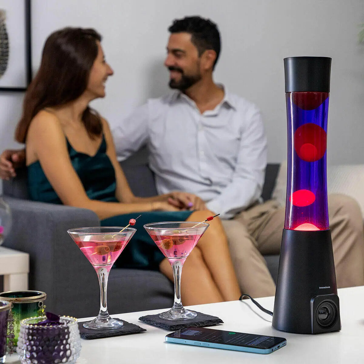 Lámpara de lava con base negra, fluido azul y burbujas rojas, junto a una pareja en un salón con copas de cóctel.