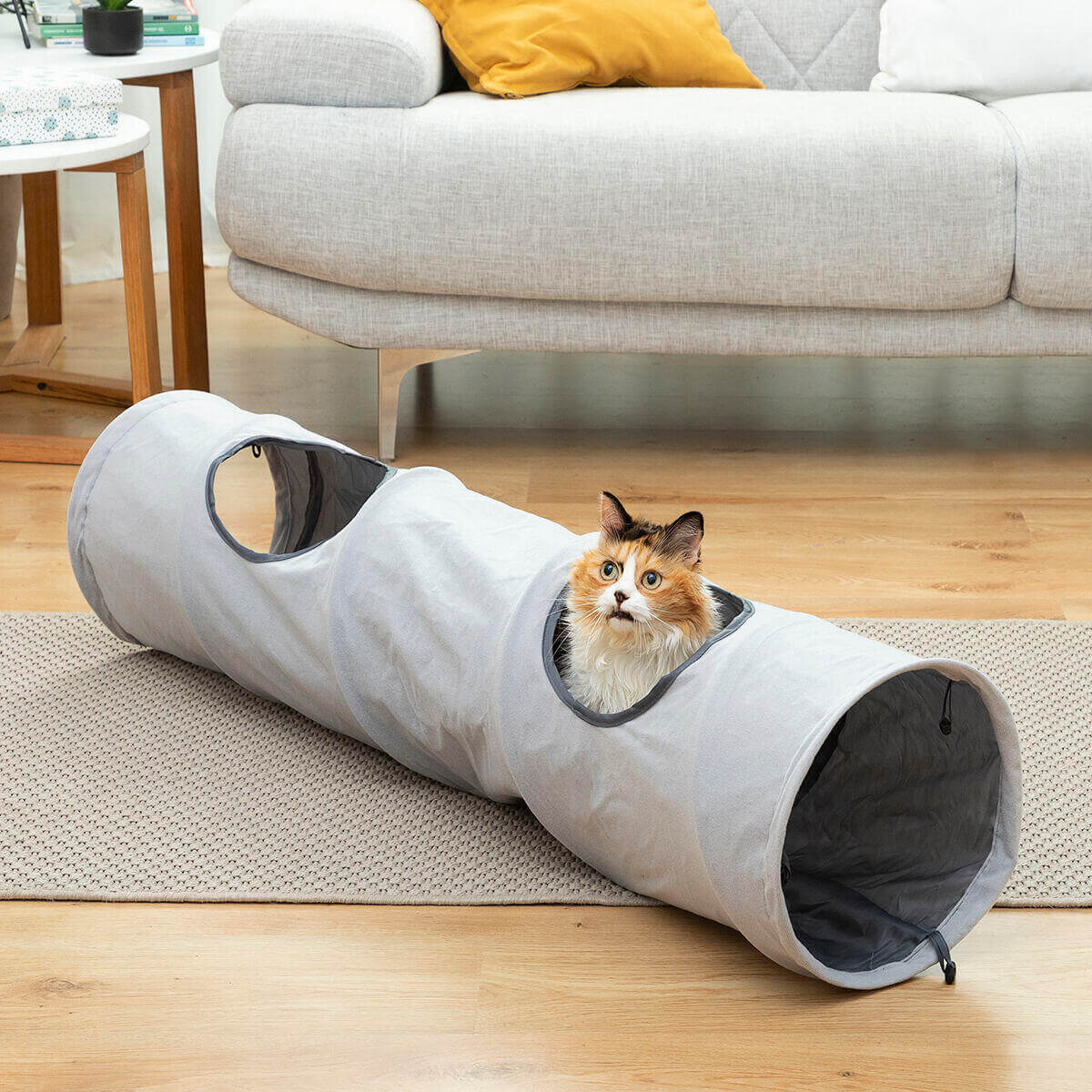 Gato tricolor asomándose por un túnel de tela gris plegable con aberturas laterales, ubicado en una sala de estar.
