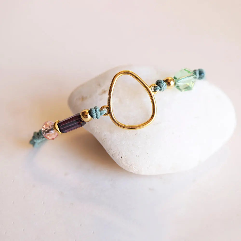 Pulsera de hilo azul, cuentas de cristal morado y verde, diseño moderno con detalle dorado en forma de gota.