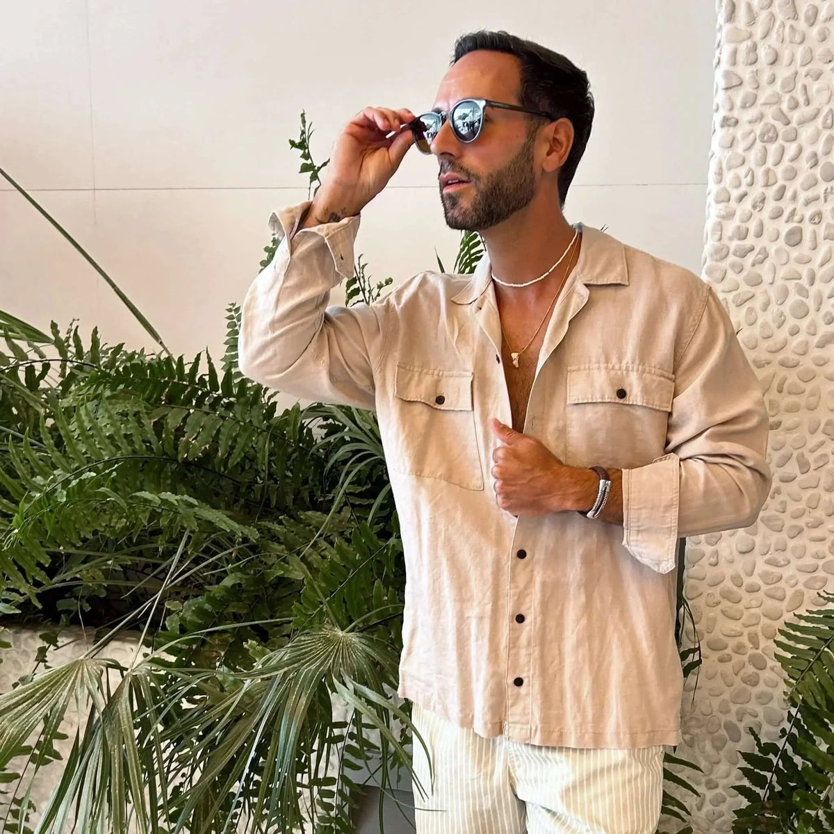 Hombre con gafas de sol redondas de montura oscura y lentes marrones, vistiendo camisa de lino beige.