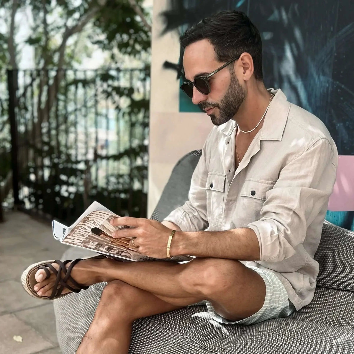 Hombre con gafas de sol redondas de montura oscura y lentes marrones, sentado al aire libre leyendo una revista.