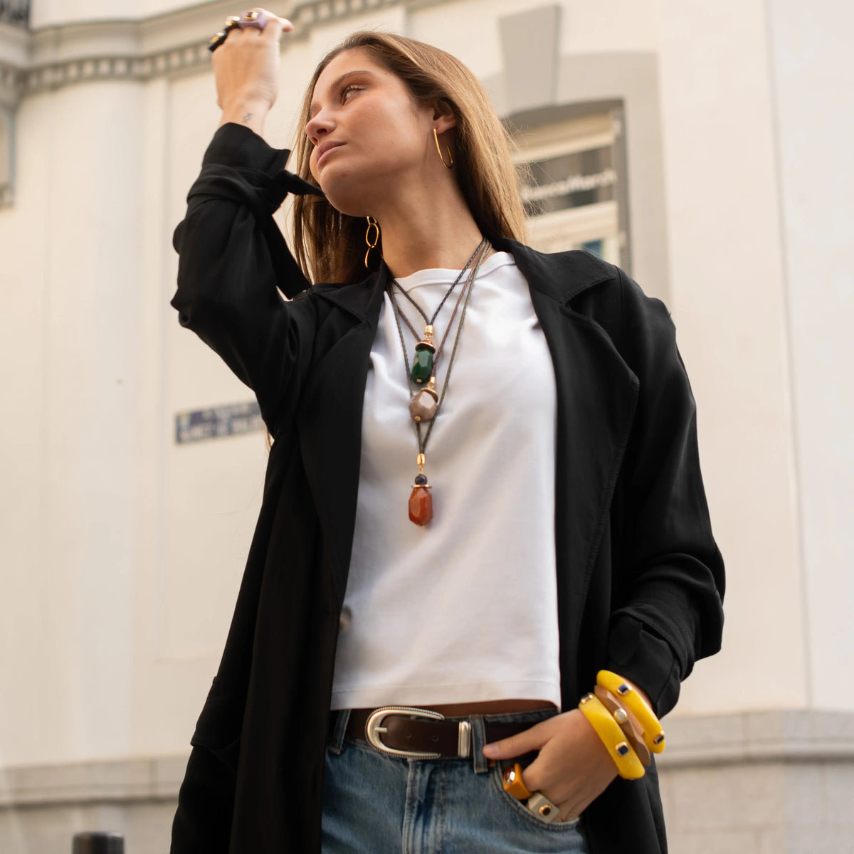 Mujer con abrigo negro, blusa blanca y jeans; accesorios de piedras y pulseras amarillas. Estilo urbano y elegante.