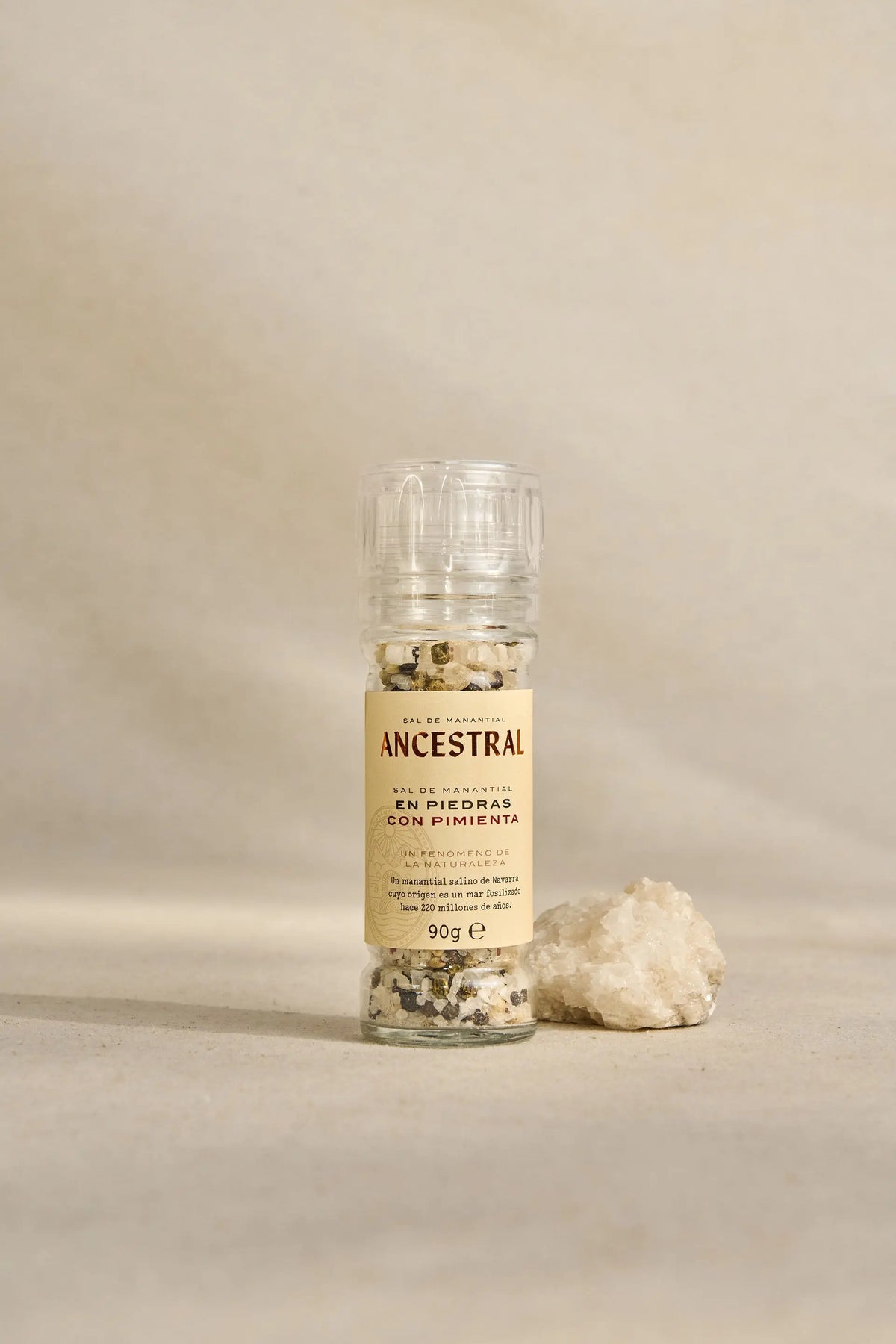 Clear glass grinder containing natural spring salt crystals and four-pepper mix, next to a raw salt rock.