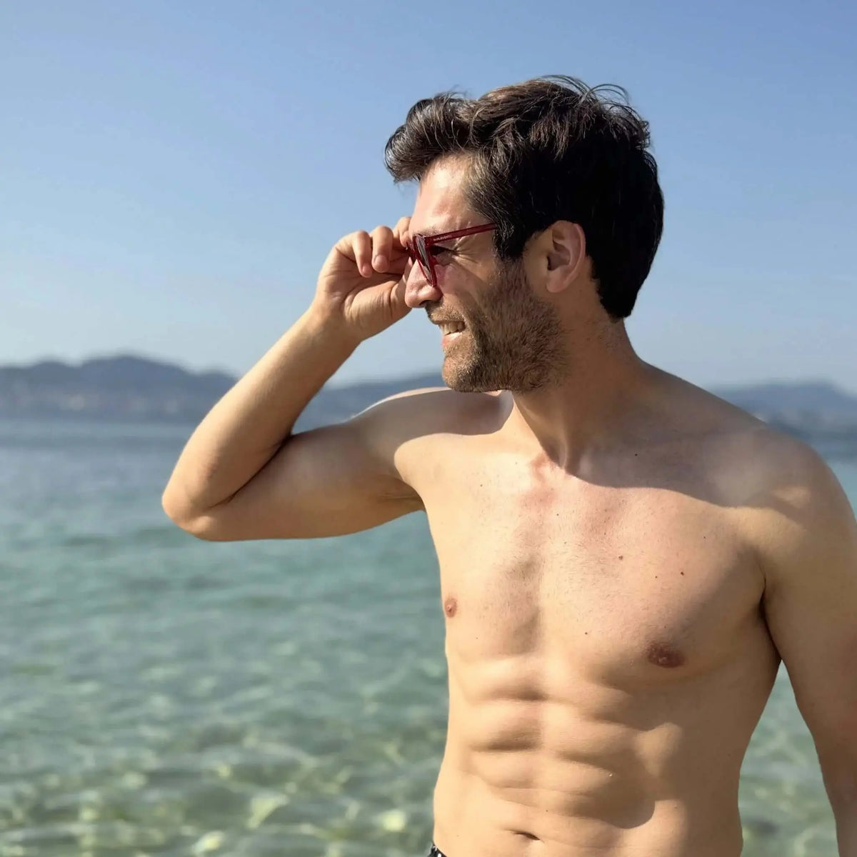 Hombre sin camiseta en la playa con gafas de sol de montura roja, estilo moderno y diseño redondo, posando frente al mar.