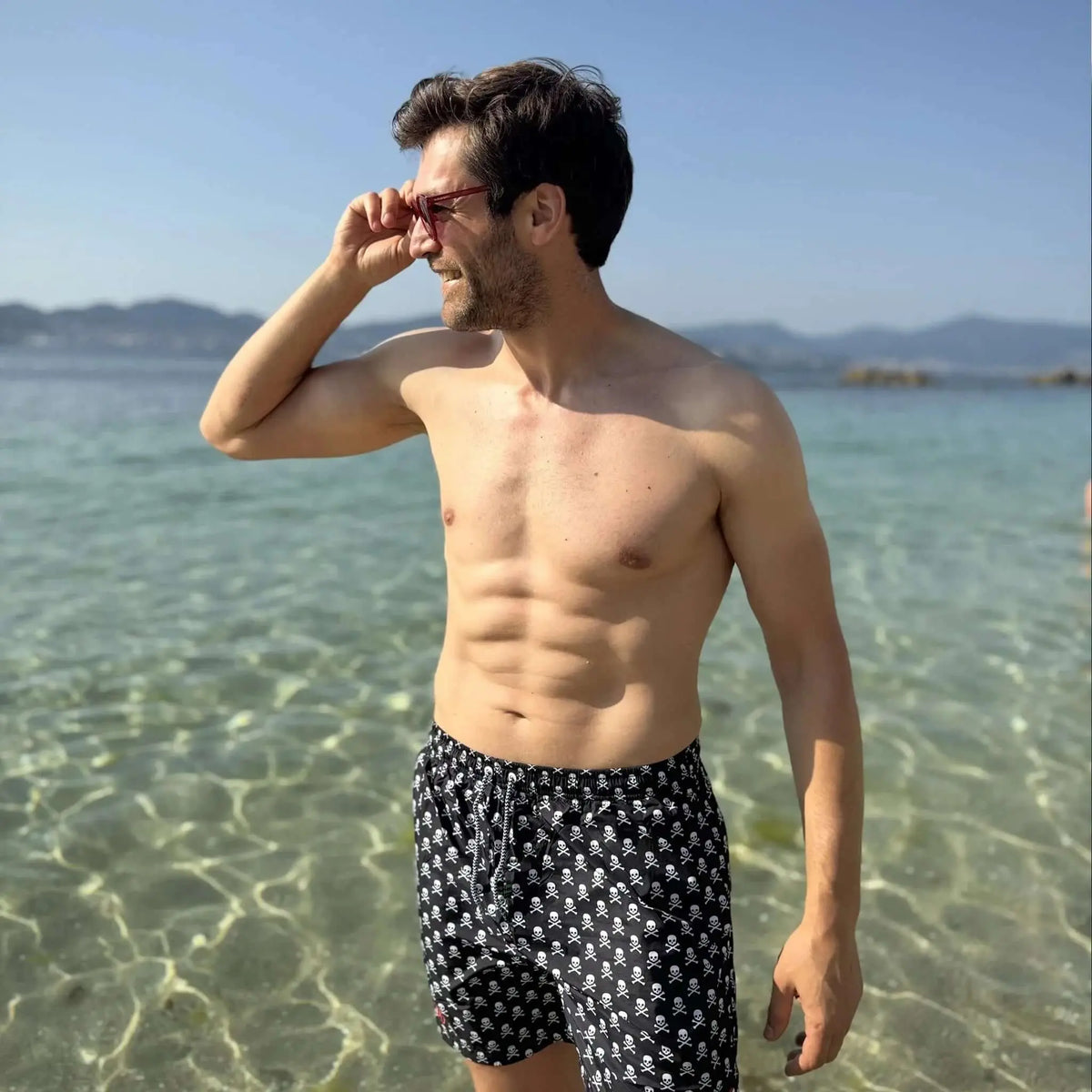 Hombre en la playa con gafas de sol redondas de montura roja, estilo casual y veraniego.
