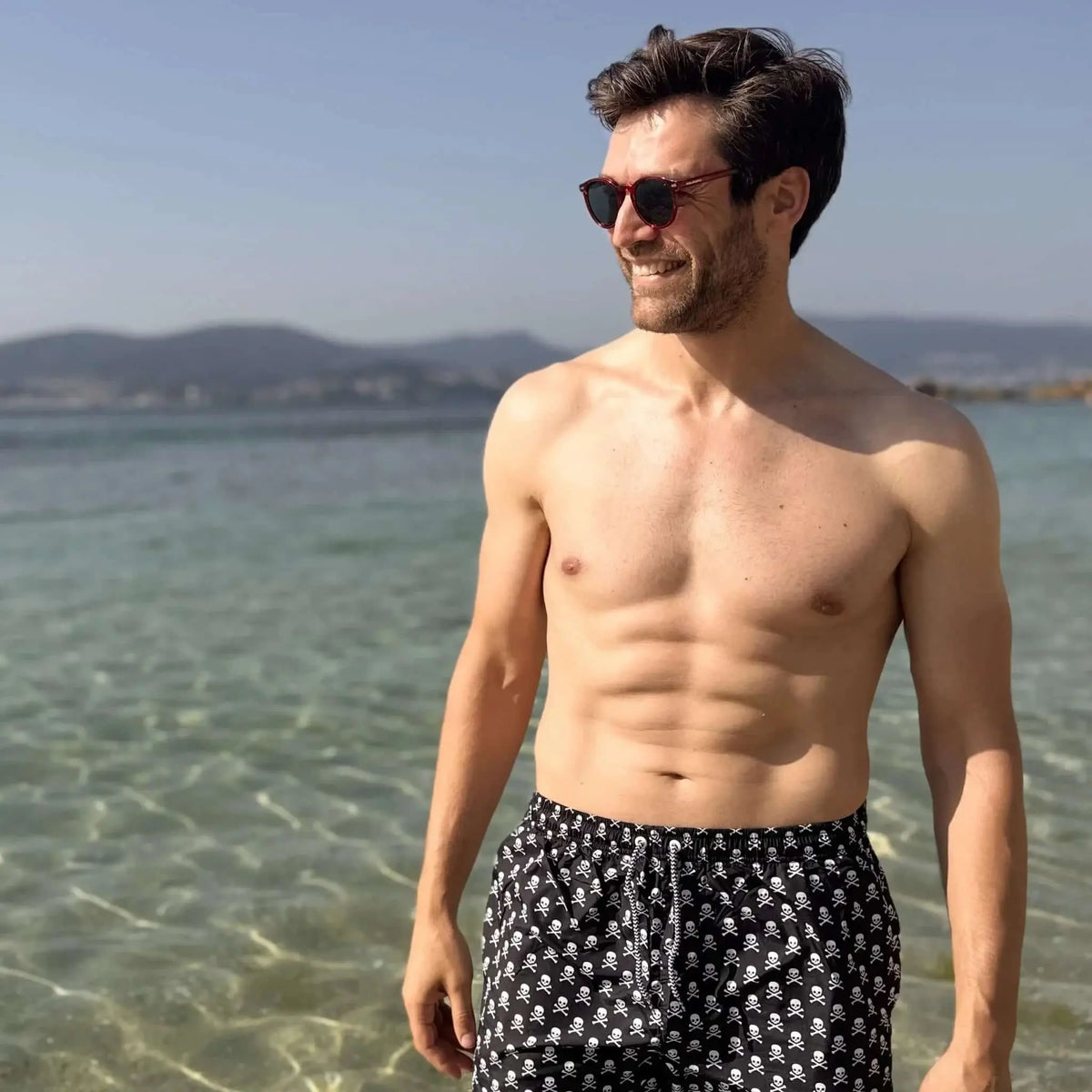 Hombre en la playa con gafas de sol redondas de montura roja y cristales oscuros, estilo veraniego y casual.