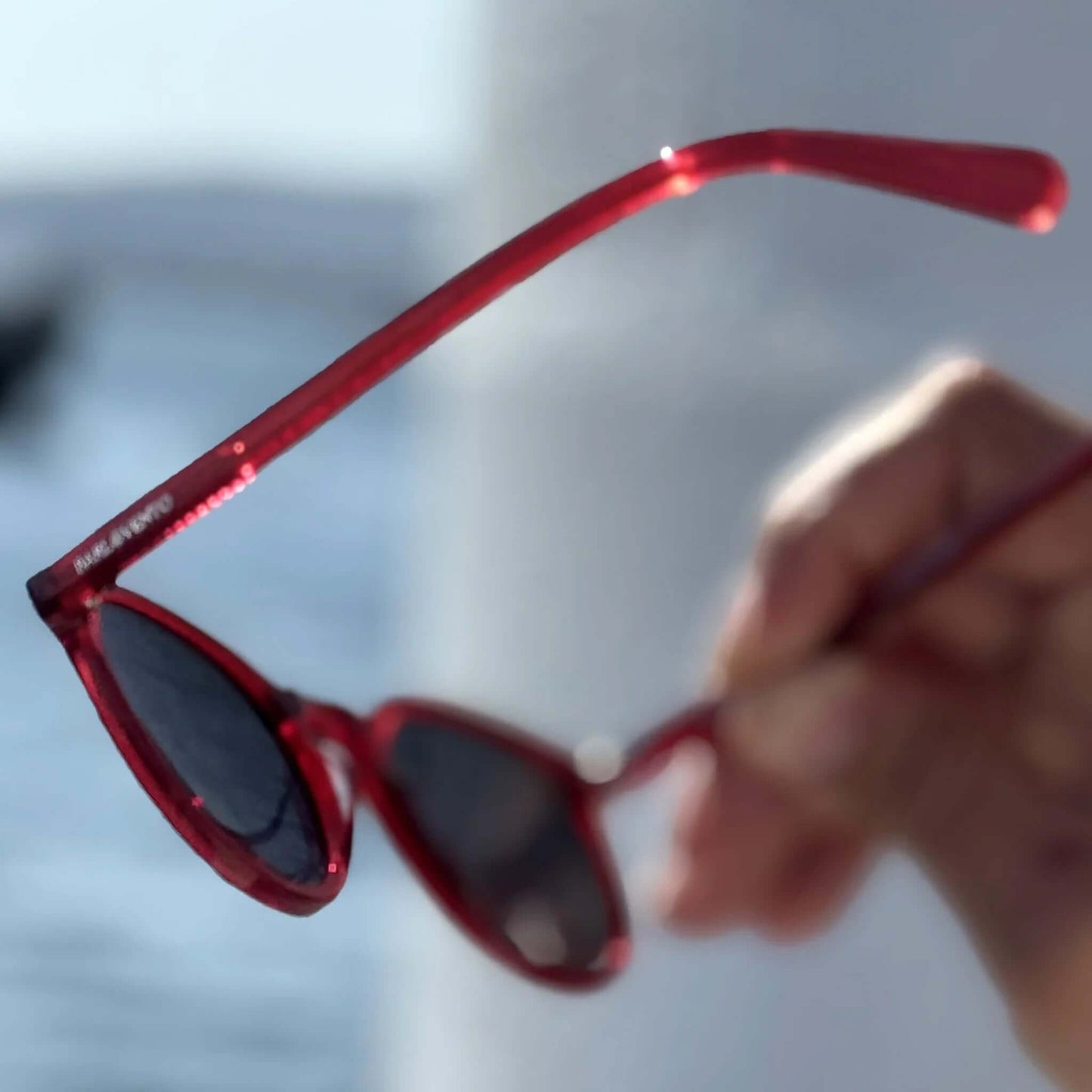 Gafas de sol redondas con montura de acetato rojo translúcido, sostenidas por una mano frente a un fondo luminoso.