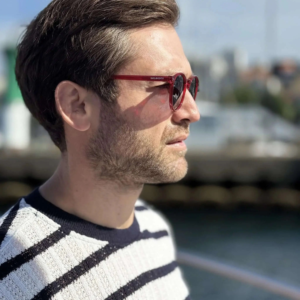 Hombre con gafas de sol redondas de montura roja translúcida y cristales oscuros, estilo casual en puerto deportivo.