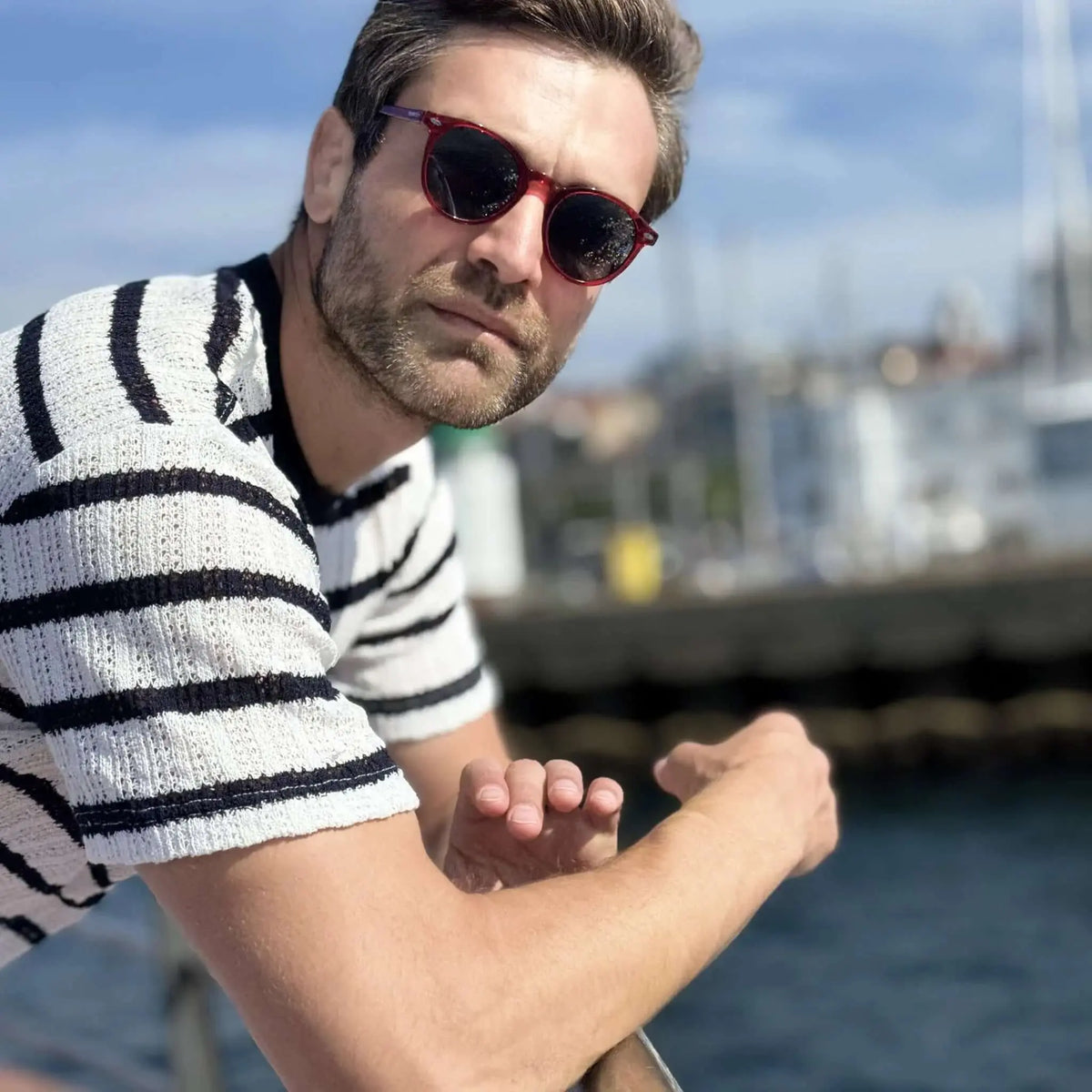 Hombre con gafas de sol redondas de montura roja translúcida y camiseta a rayas en un puerto deportivo.