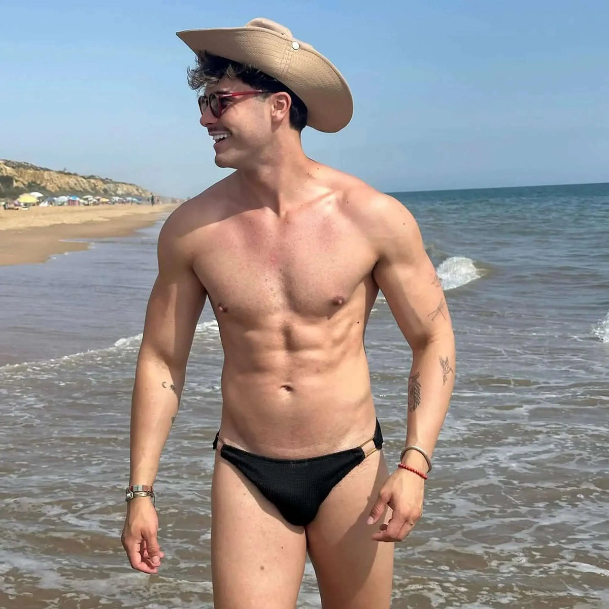 Hombre en la playa con gafas de sol redondas de montura roja, sombrero y bañador negro, estilo veraniego.