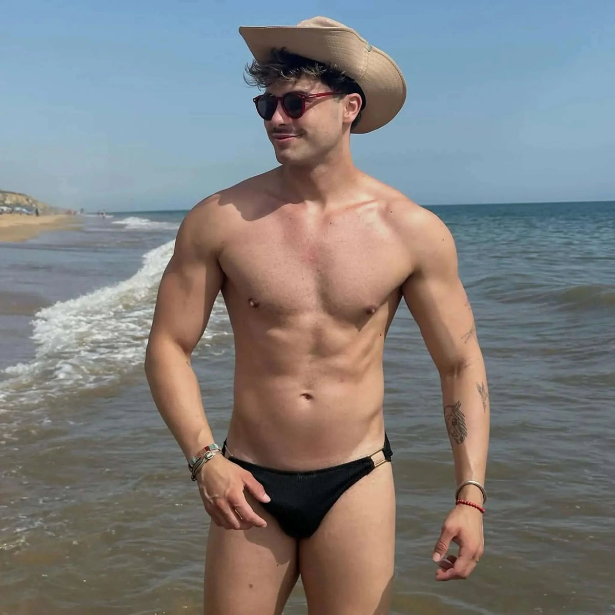 Hombre en la playa con gafas de sol redondas de montura roja y lentes oscuras, estilo veraniego y casual.