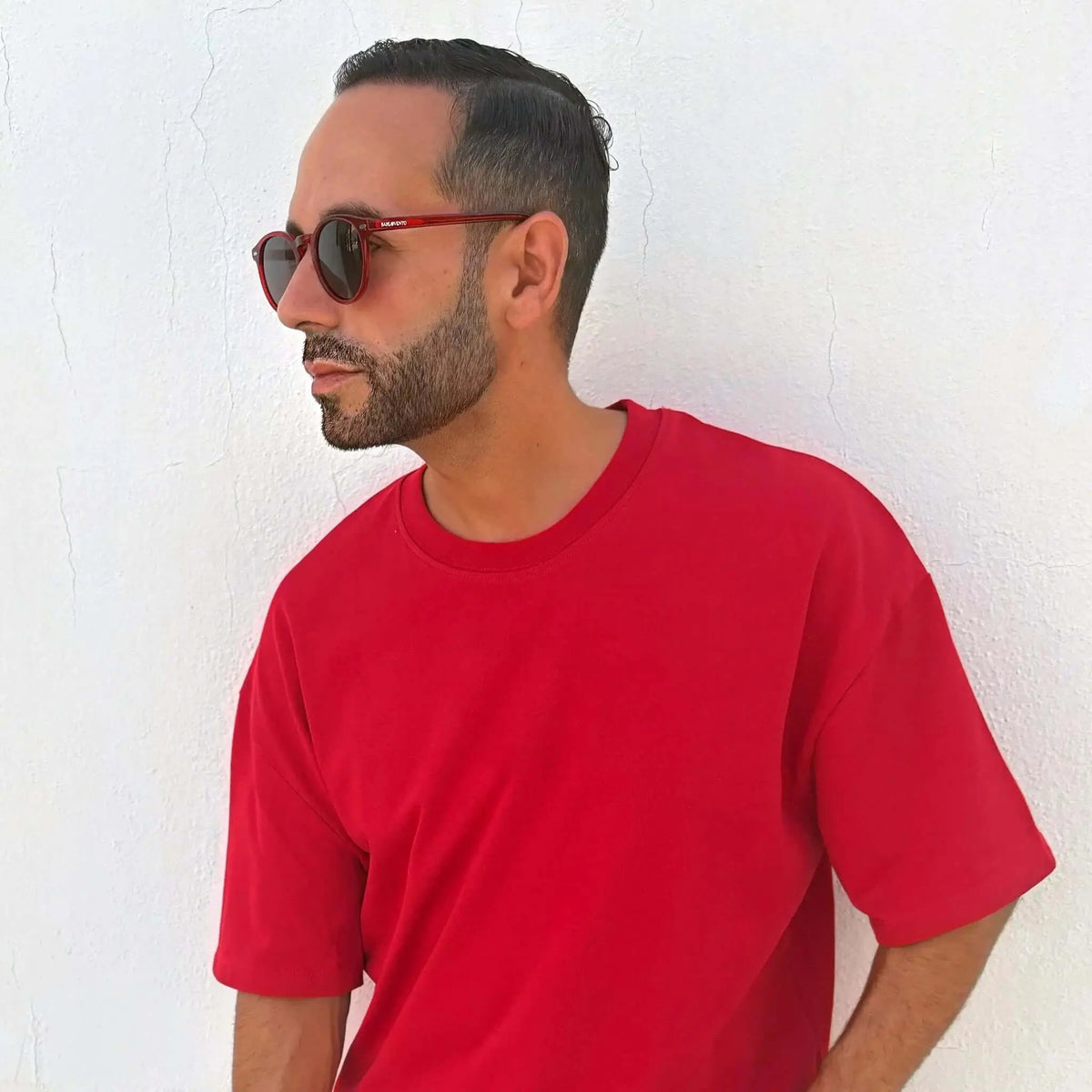 Hombre con camiseta roja y gafas de sol redondas de montura roja translúcida contra pared blanca. Estilo moderno.
