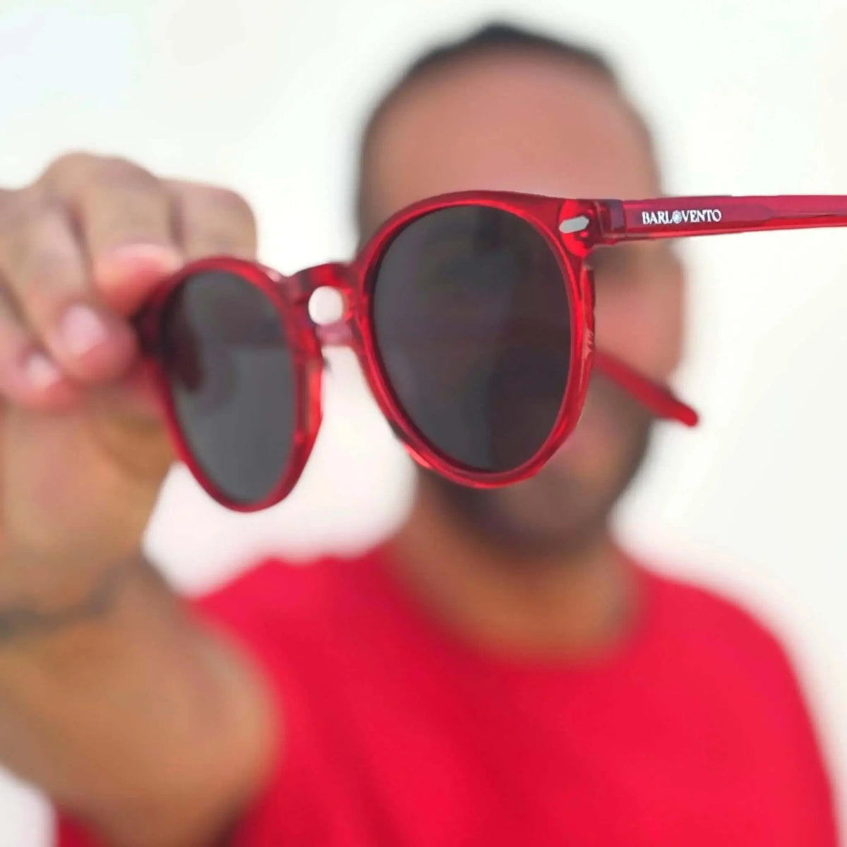 Gafas de sol redondas con montura de acetato rojo translúcido y cristales oscuros sostenidas por un hombre de rojo.
