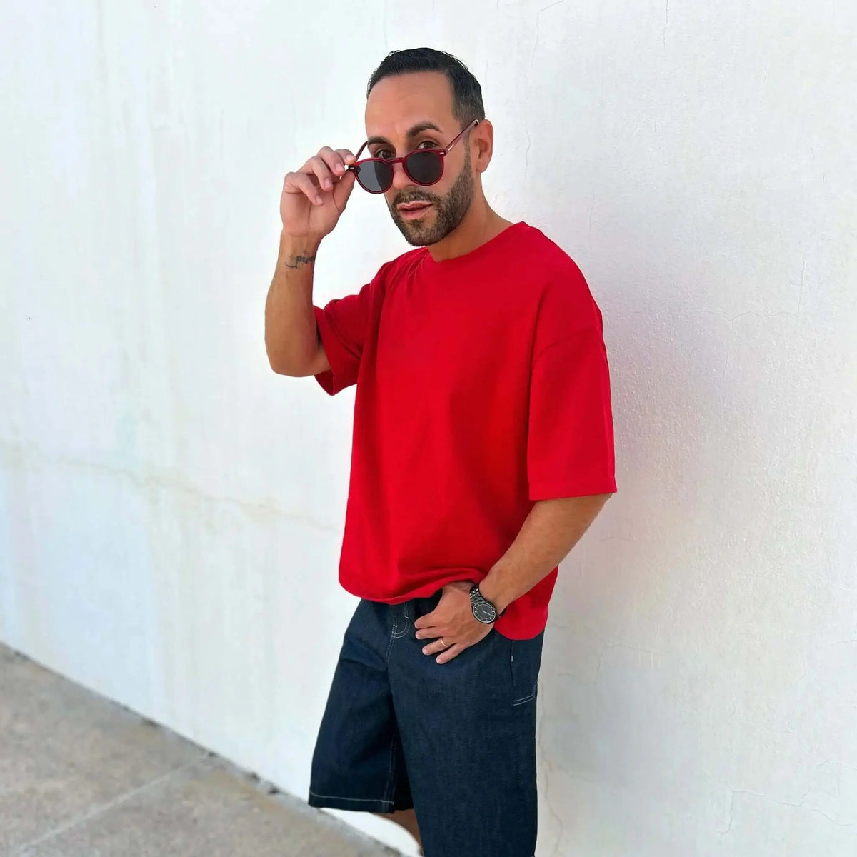 Hombre con gafas de sol redondas de montura roja, camiseta roja y bermudas vaqueras posando contra una pared blanca.