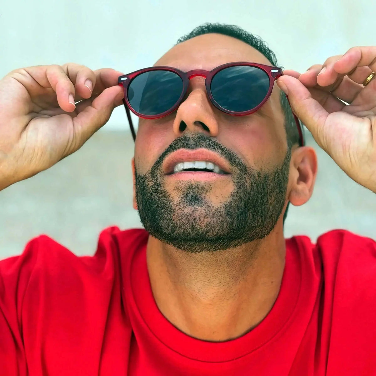 Hombre con camiseta roja y gafas de sol redondas de montura roja transparente y cristales oscuros. Estilo casual.