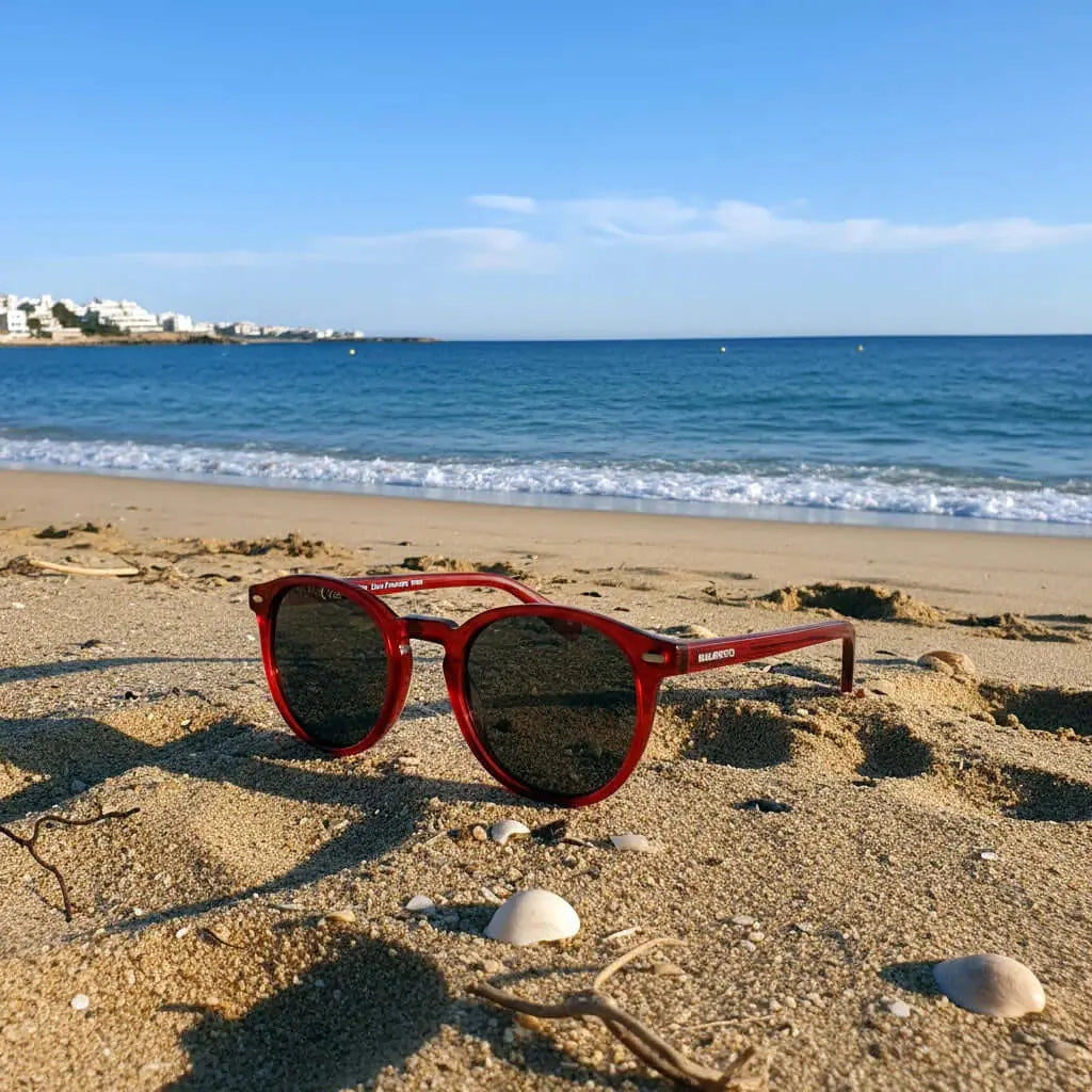 Gafas de sol redondas con montura roja traslúcida y cristales oscuros sobre la arena de una playa soleada.