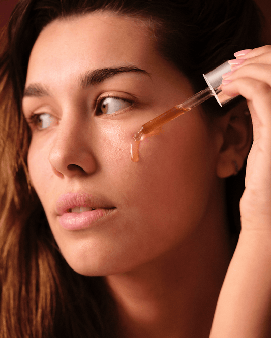 Mujer aplicando sérum facial dorado Banbu con gotero sobre su mejilla. Textura fluida en un ambiente cálido y natural.