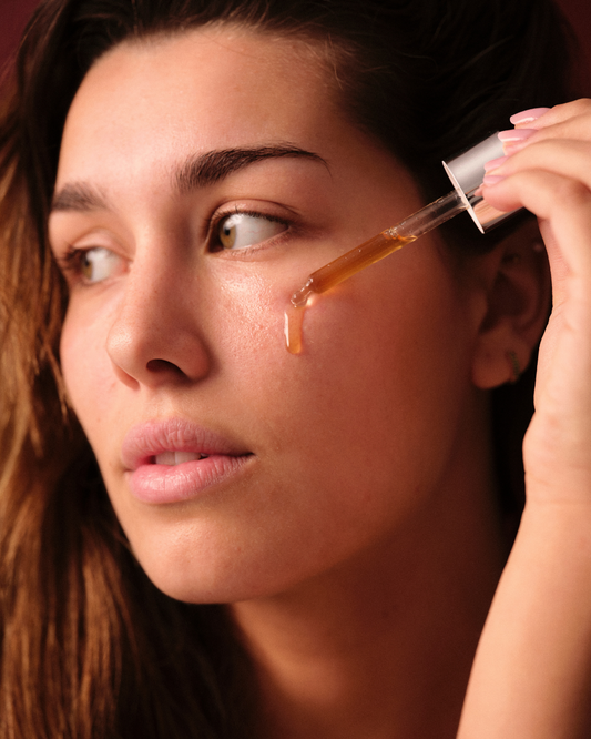 Mujer aplicando sérum facial dorado Banbu con gotero sobre su mejilla. Textura fluida en un ambiente cálido y natural.