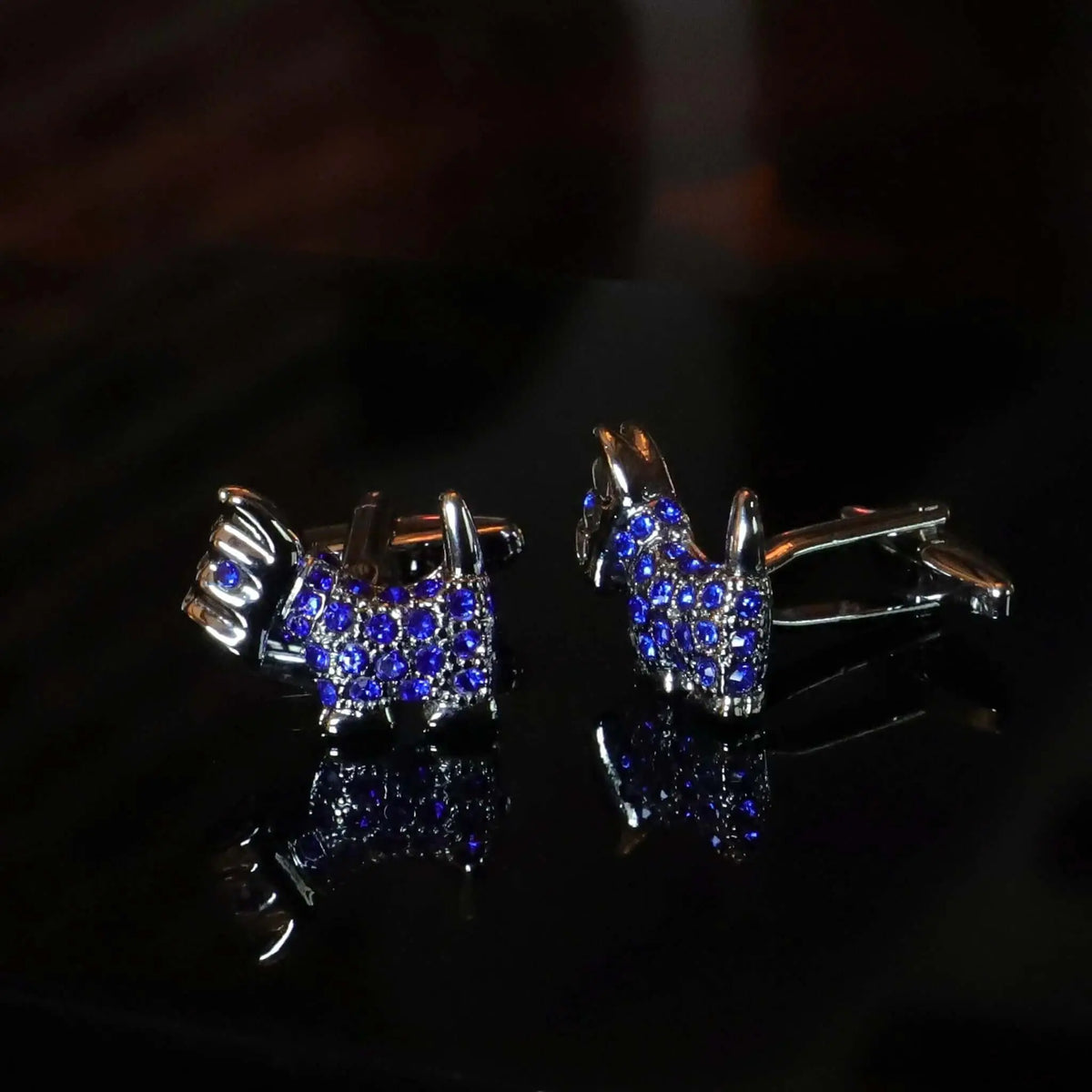 Gemelos plateados con forma de perro terrier escocés, decorados con pequeñas gemas azules sobre fondo negro.