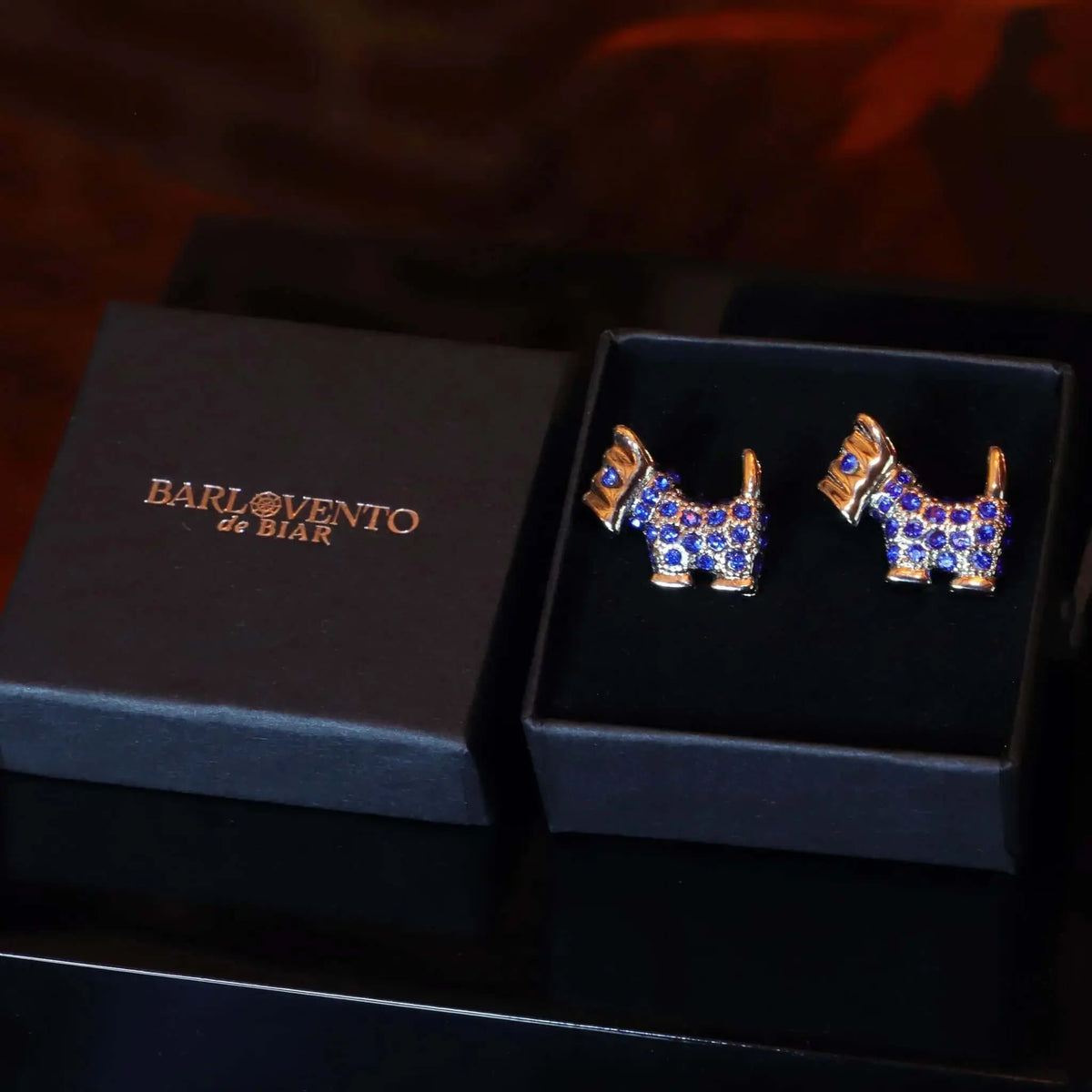 Gemelos plateados con forma de perro Terrier escocés, incrustados con brillantes gemas azules en una caja negra.