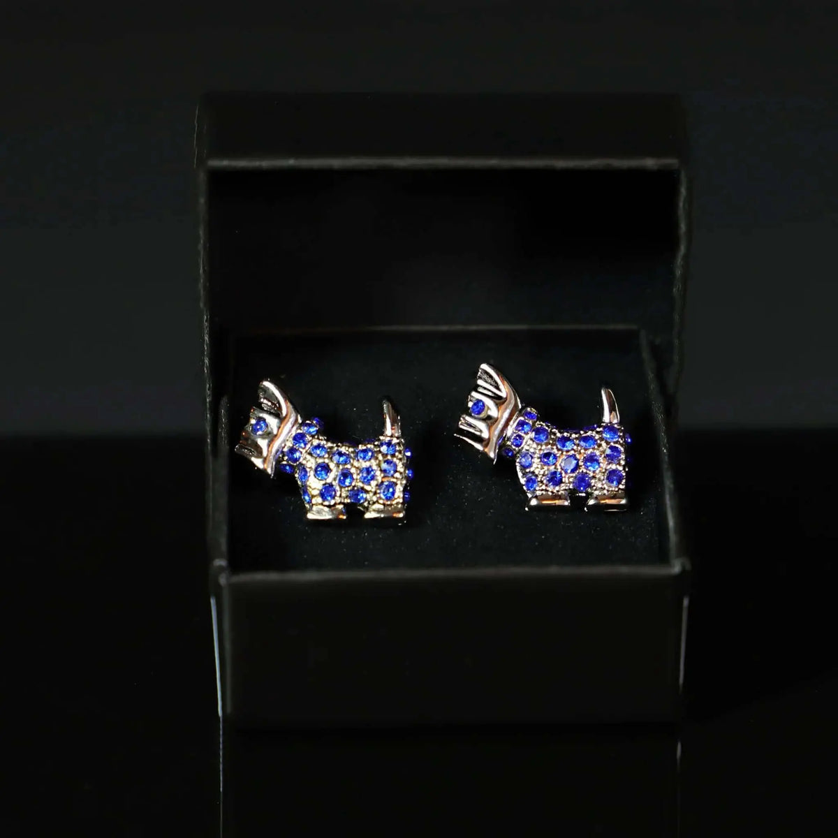 Gemelos plateados con forma de perro Terrier, decorados con pequeñas piedras azules brillantes, dentro de caja negra.