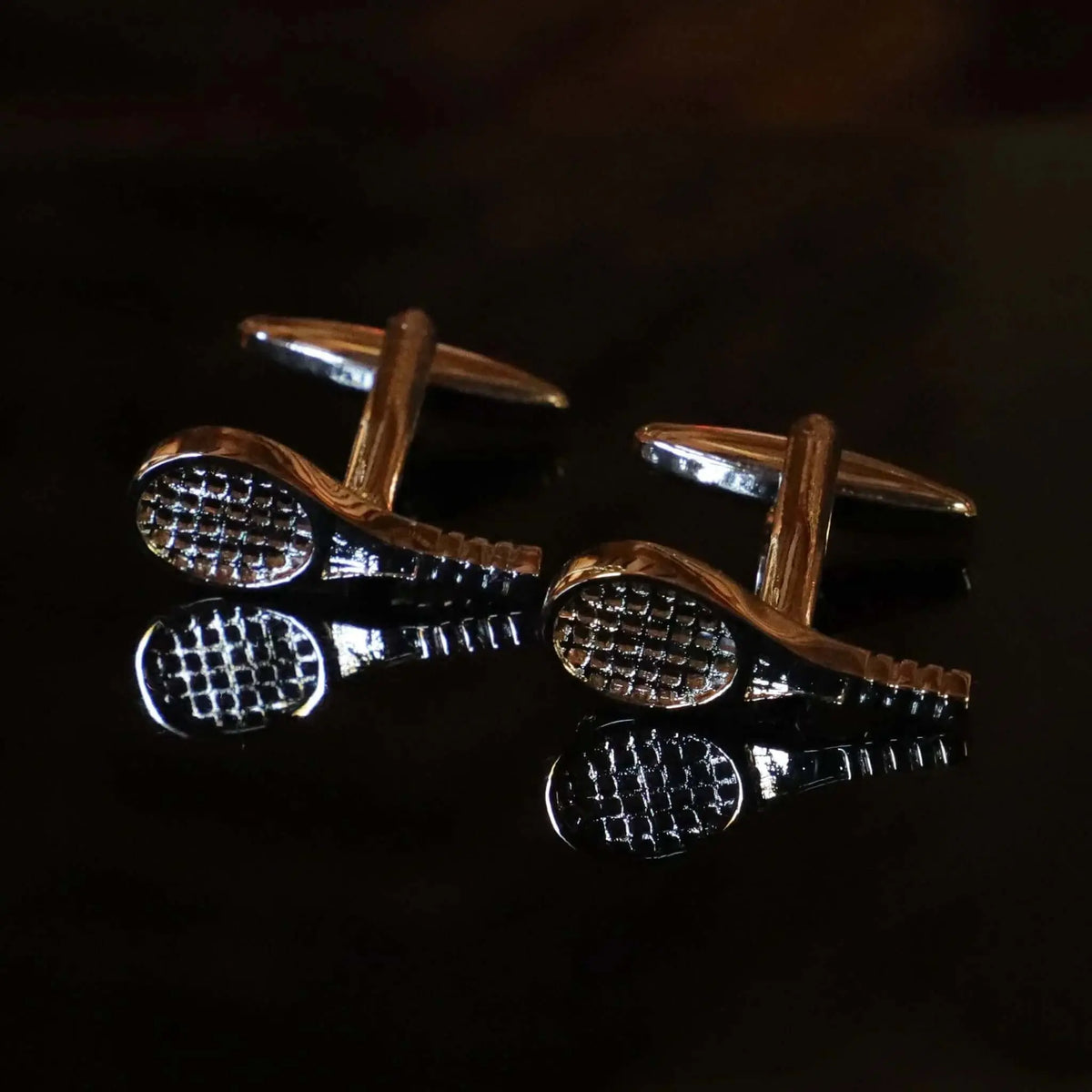 Gemelos plateados con forma de raqueta de tenis, acabado metálico brillante sobre fondo oscuro con reflejo.