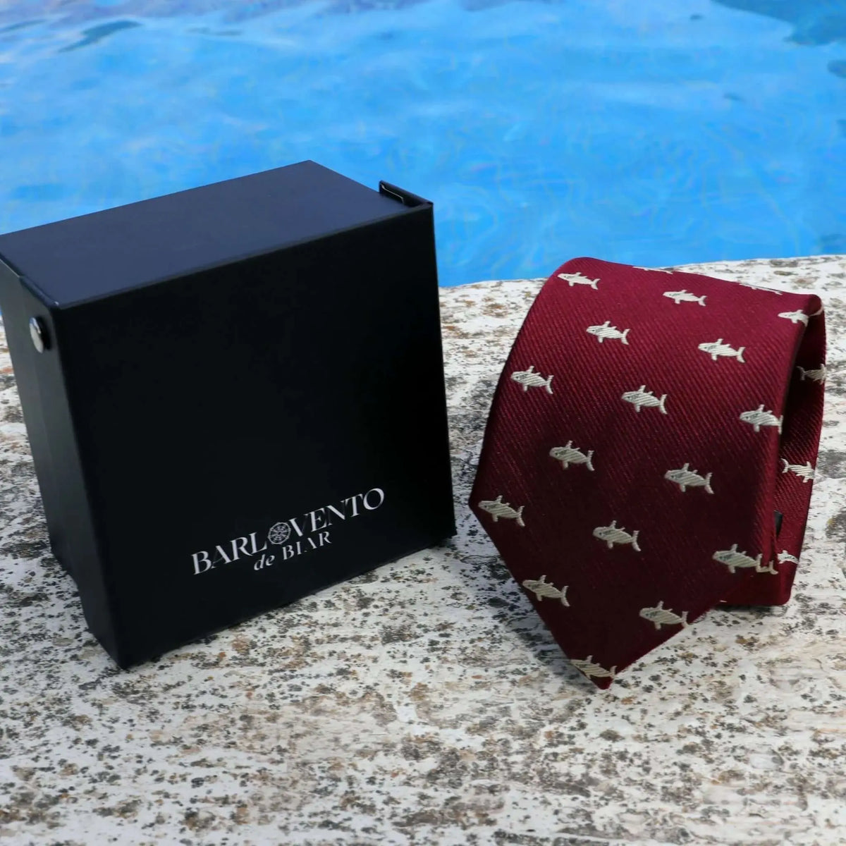 Corbata de seda color burdeos con estampado de tiburones pequeños y caja negra junto a una piscina.