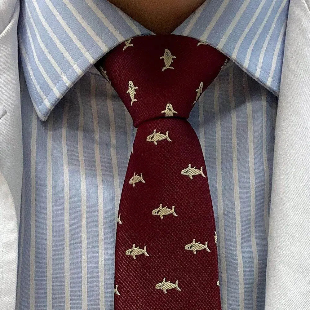 Corbata de diseño en color burdeos con estampado de pequeños tiburones beige sobre camisa de rayas azul y blanca.