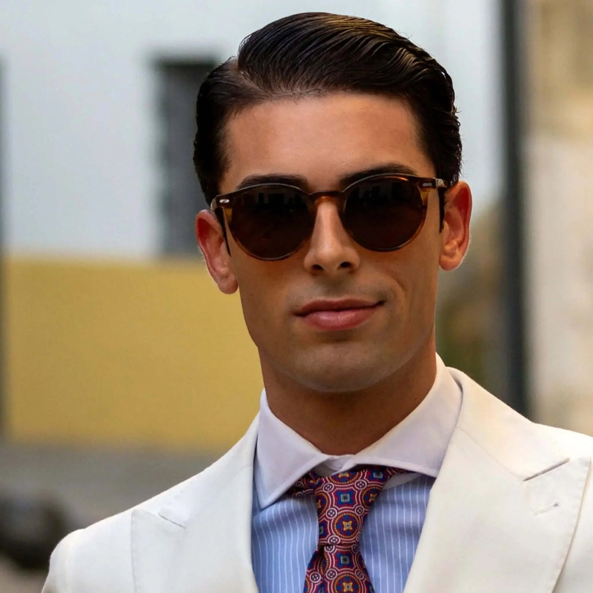 Hombre con traje blanco y gafas de sol redondas de montura naranja cristal y lentes marrones de estilo elegante.
