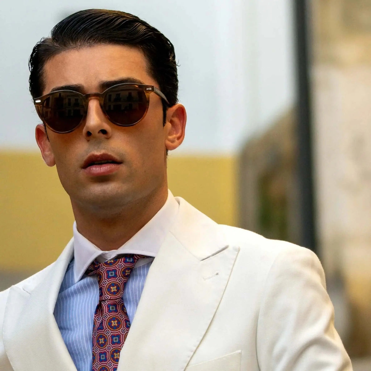 Hombre elegante con gafas de sol redondas de montura naranja cristal y lentes marrones, traje blanco y corbata.