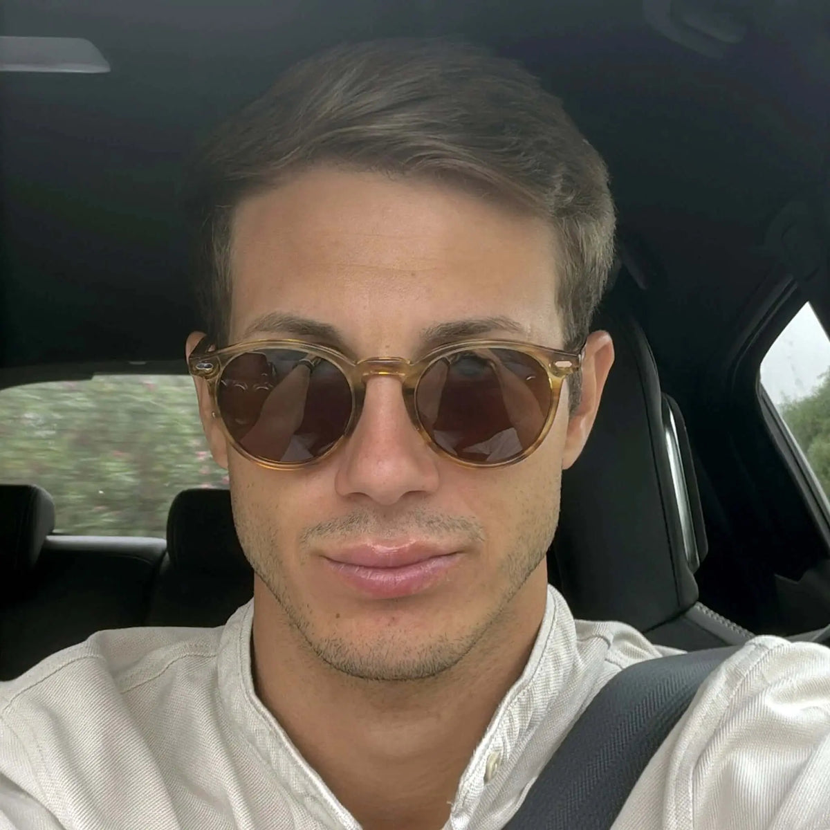 Hombre con gafas de sol redondas de montura naranja translúcida y lentes marrones, sentado en el coche con camisa beige.