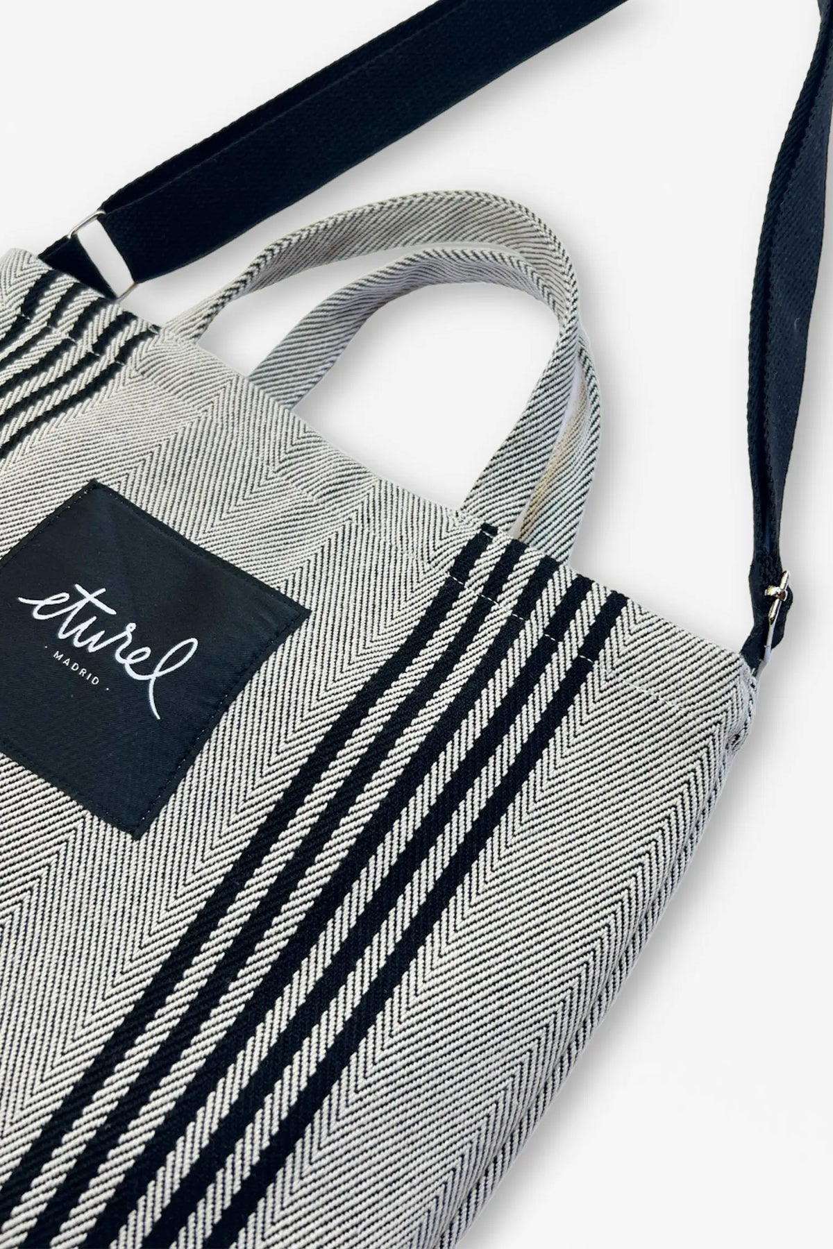 Bolso bandolera de sarga en blanco y negro con patrón de espiga, asas cortas y correa ajustable sobre fondo neutro.