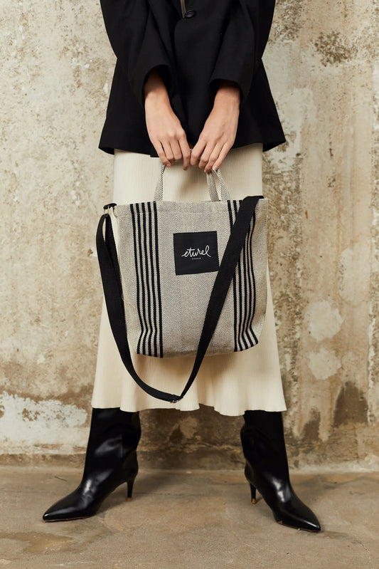 Black and cream striped twill tote bag with top handles, a black crossbody strap, and a square brand logo patch.