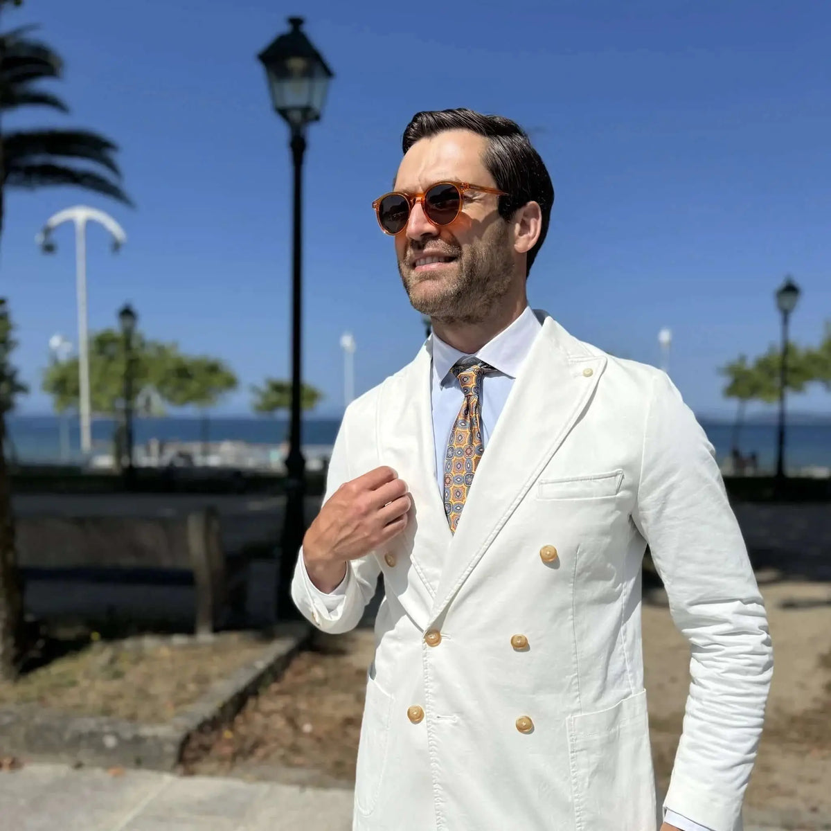 Hombre con traje blanco y gafas de sol redondas de acetato color naranja cristal, estilo elegante en paseo marítimo.