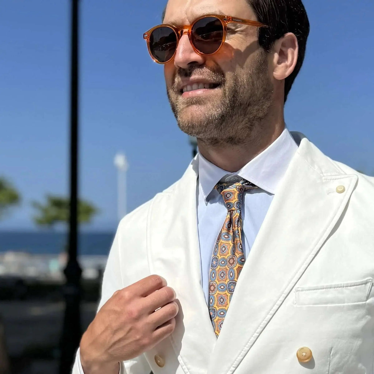 Hombre con traje blanco y gafas de sol de montura redonda color naranja transparente en un entorno soleado.