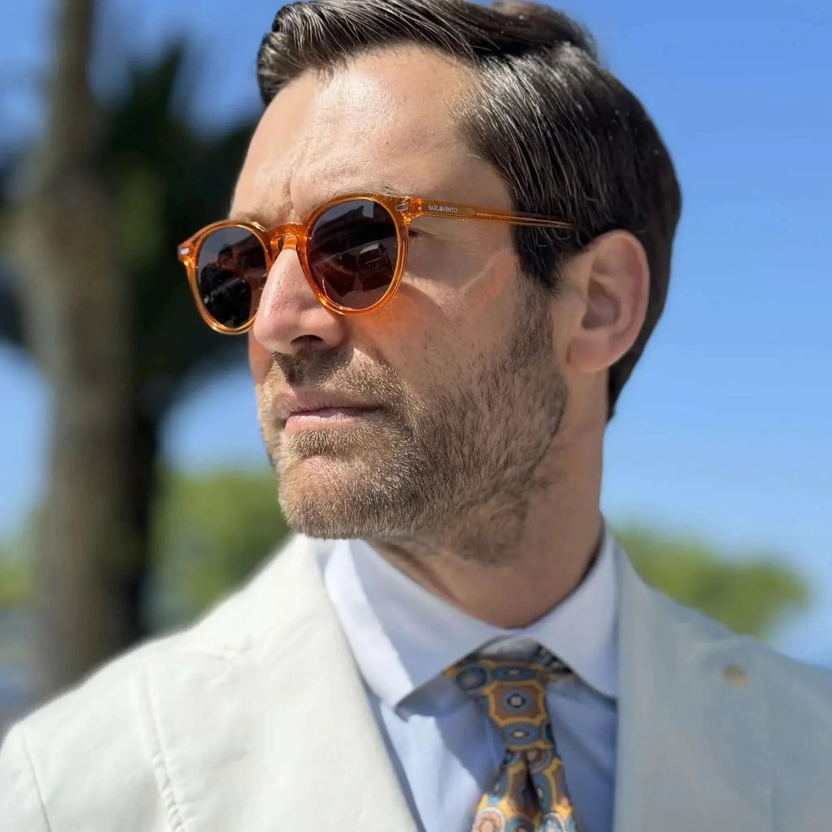 Hombre con traje y gafas de sol de pasta redondas color naranja translúcido, estilo elegante en exteriores.