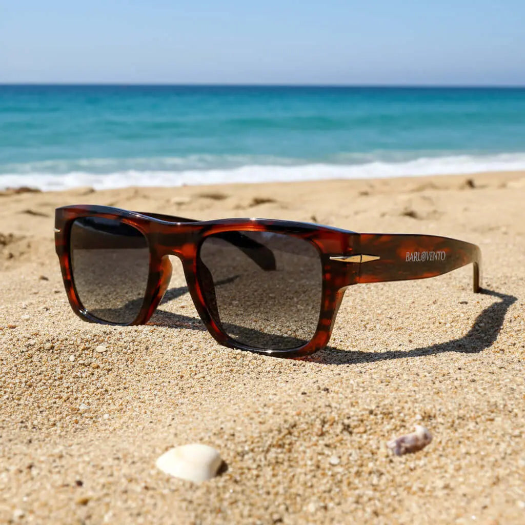 Gafas de sol de carey con lentes oscuras sobre la arena de una playa con el mar al fondo.