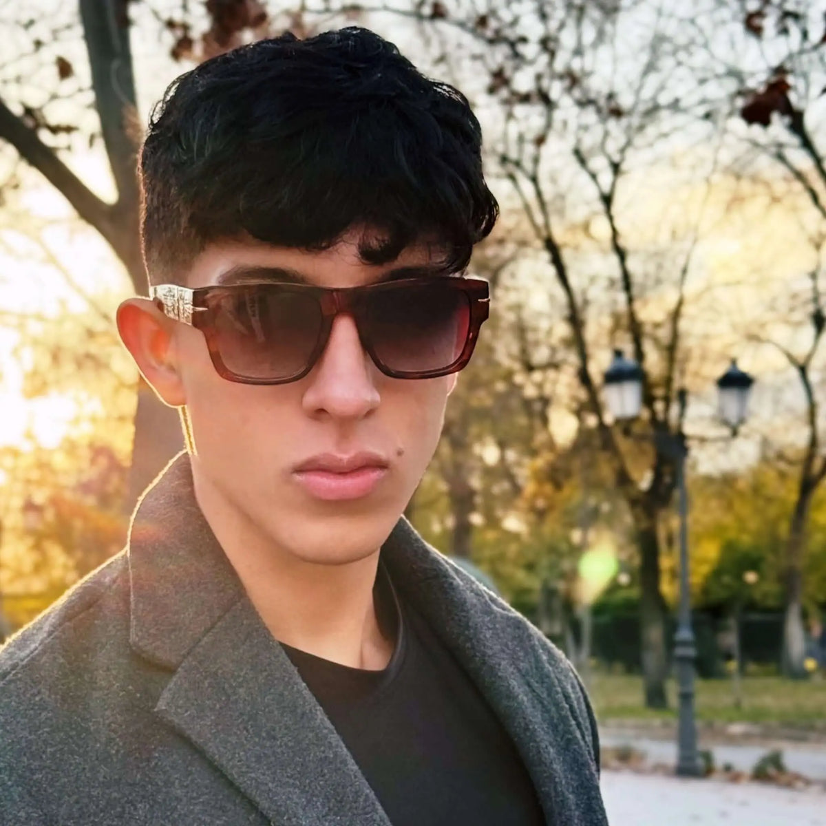 Joven con abrigo gris y gafas de sol rectangulares estilo carey con lentes azules en un parque al atardecer.