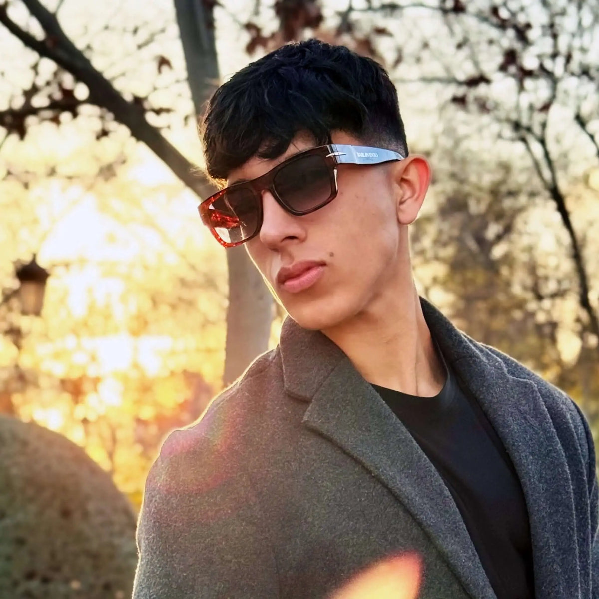 Joven con abrigo y gafas de sol de carey estilo cuadrado con lentes degradadas y protección UV en un parque al atardecer.