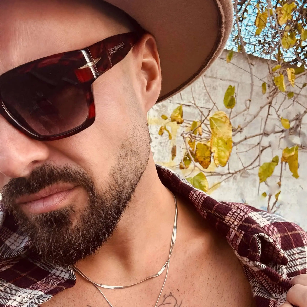 Hombre con sombrero y camisa de cuadros usa gafas de sol de carey con lentes azules, estilo elegante y moderno.