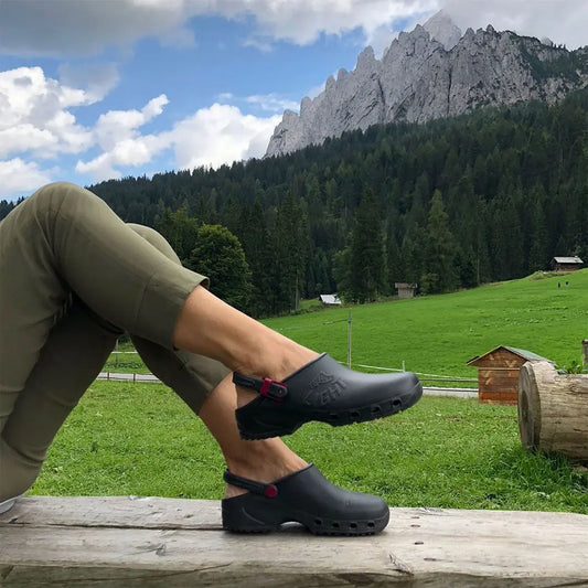Black slip-on clogs with a heel strap and side cutouts, worn against a scenic mountain landscape.