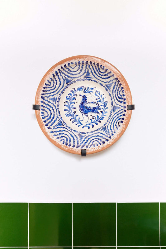 Soporte de pared de hierro negro sosteniendo un lebrillo de cerámica decorado con motivos en azul sobre fondo blanco.