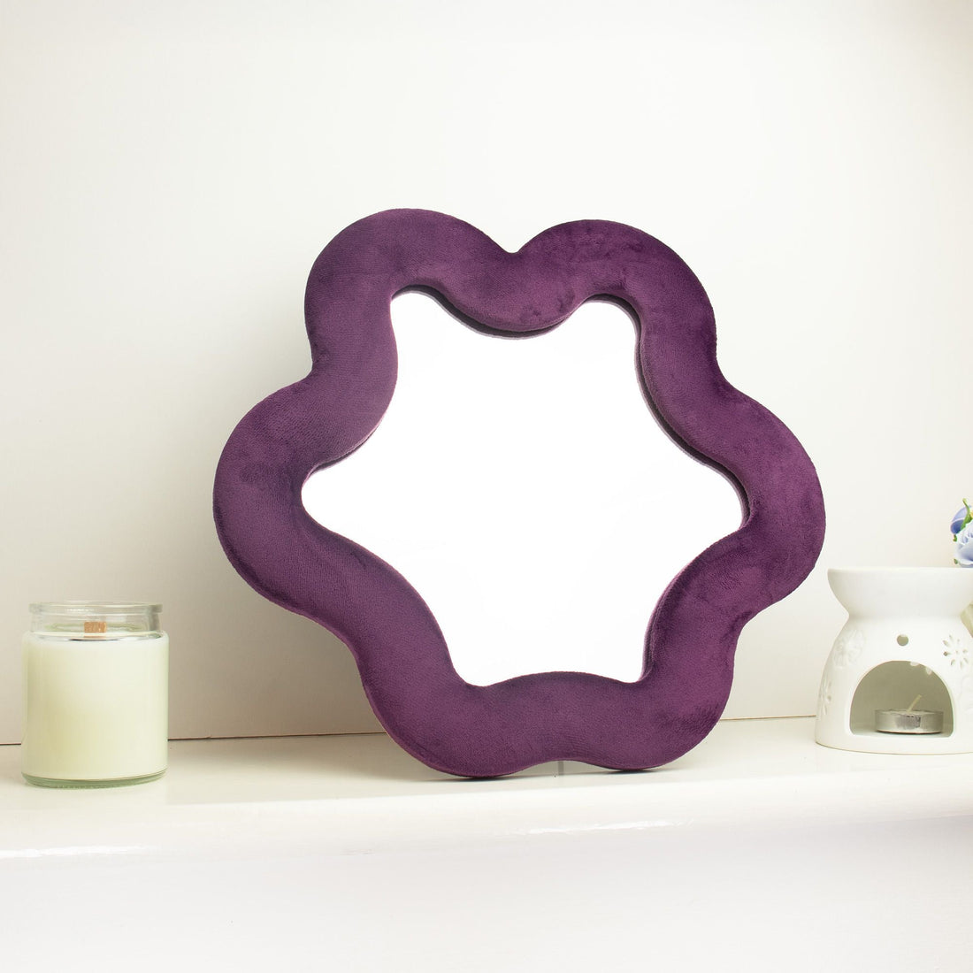 Espejo estrella malva ondulado con marco de terciopelo sobre estantería blanca junto a vela decorativa