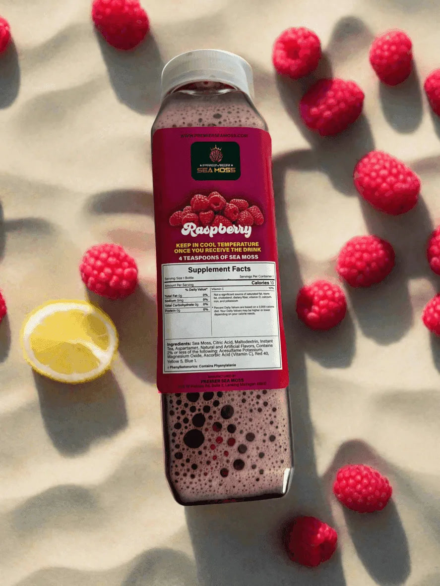 12 oz bottle of pink raspberry sea moss lemonade surrounded by fresh raspberries and a lemon slice on sand.