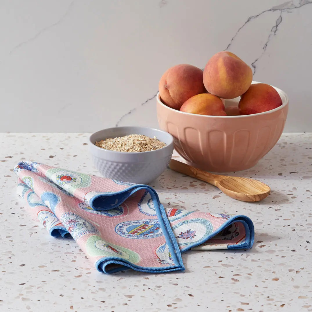 Microfiber towel with a pink background and vintage plate pattern, featuring a blue stitched edge on a stone counter.