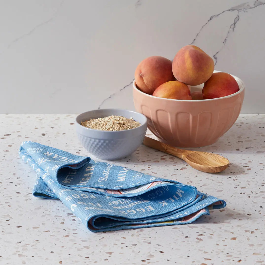Blue microfiber towel with baking-themed text print, displayed on a speckled countertop next to fruit and oats.