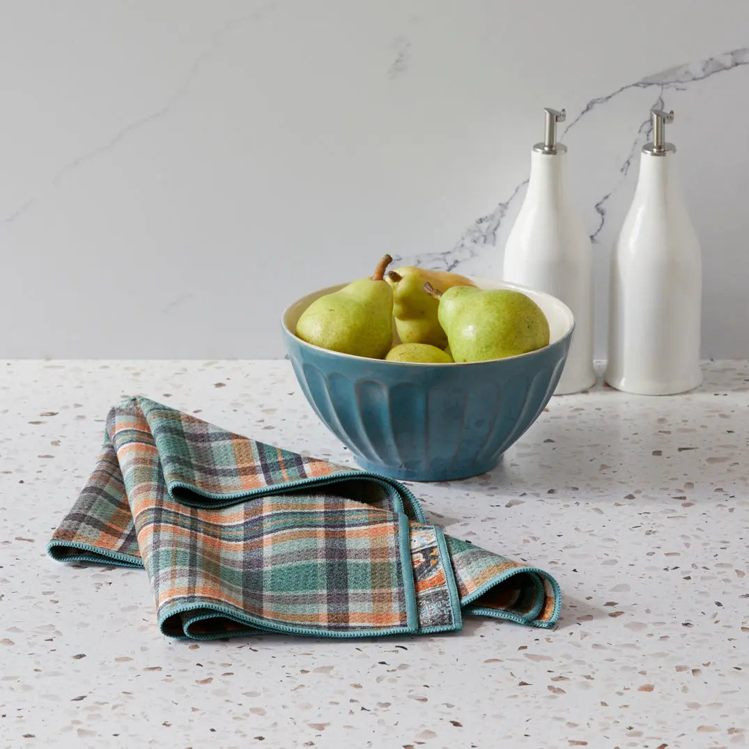 Microfiber towel with a blue, orange, and tan plaid pattern resting on a countertop next to a blue bowl of pears.