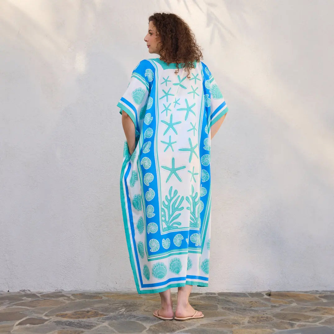 Woman wearing a white caftan featuring turquoise starfish, coral, and seashell prints with blue geometric borders.